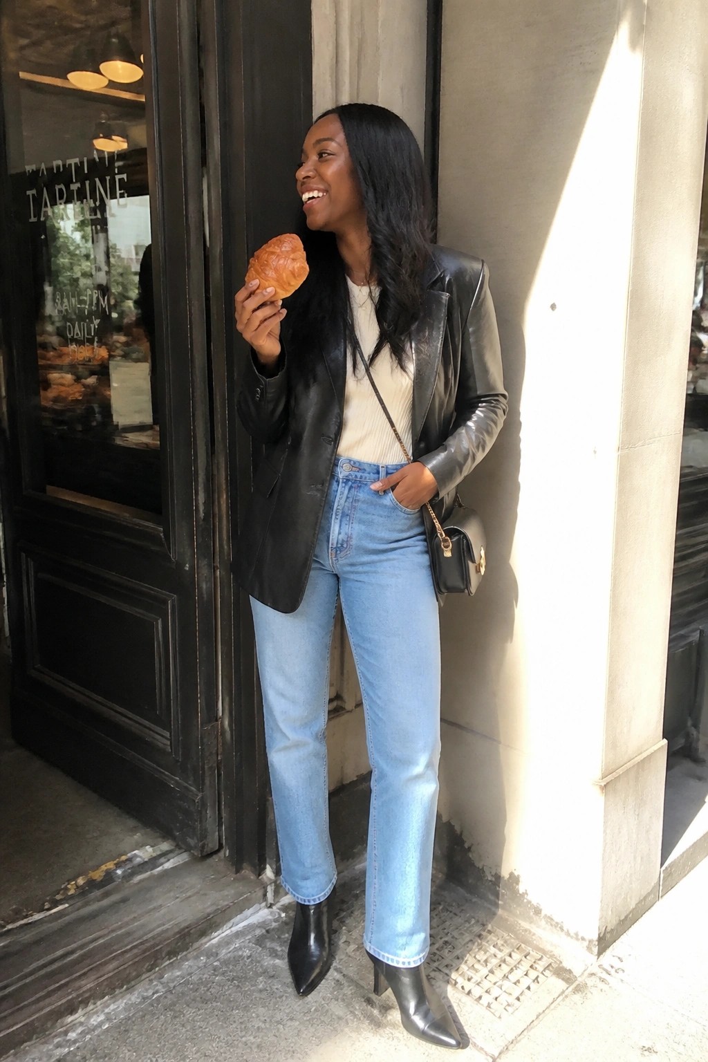 Smiling woman in black leather jacket holds croissant outside bakery in sunlight.