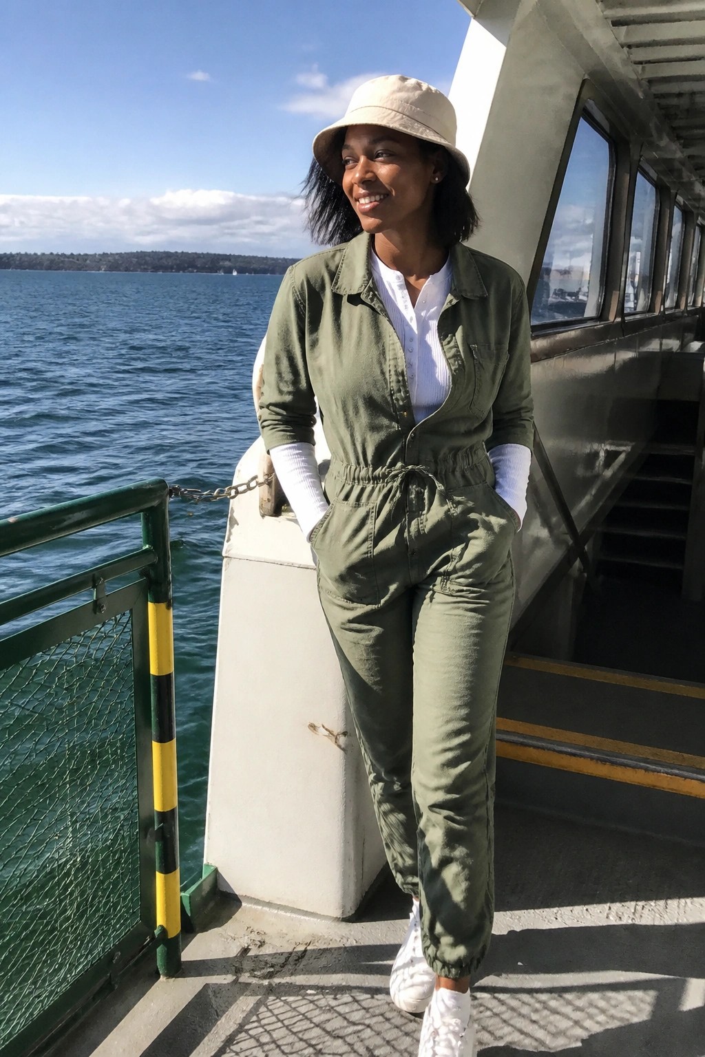 Smiling woman in green jumpsuit and beige hat on sunny ferry deck by water.