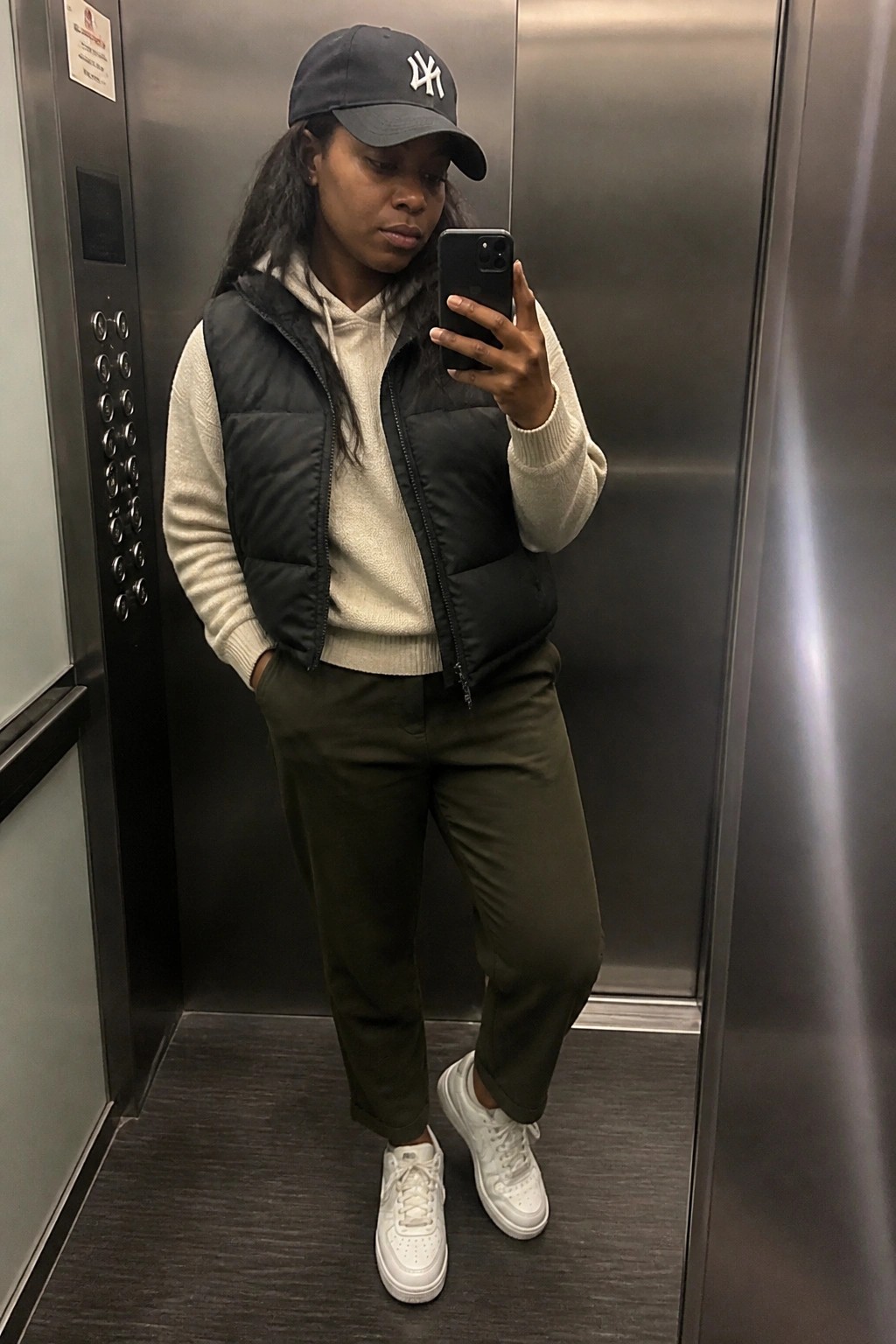 Woman in black puffer vest and NY cap taking mirror selfie in elevator