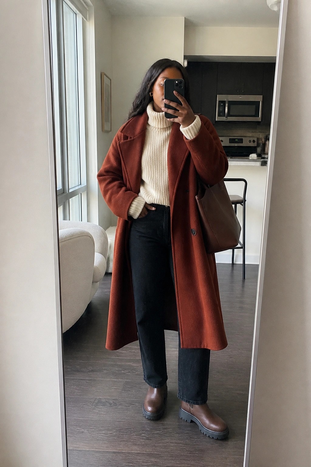 Woman in rust coat over cream turtleneck taking mirror selfie in apartment.