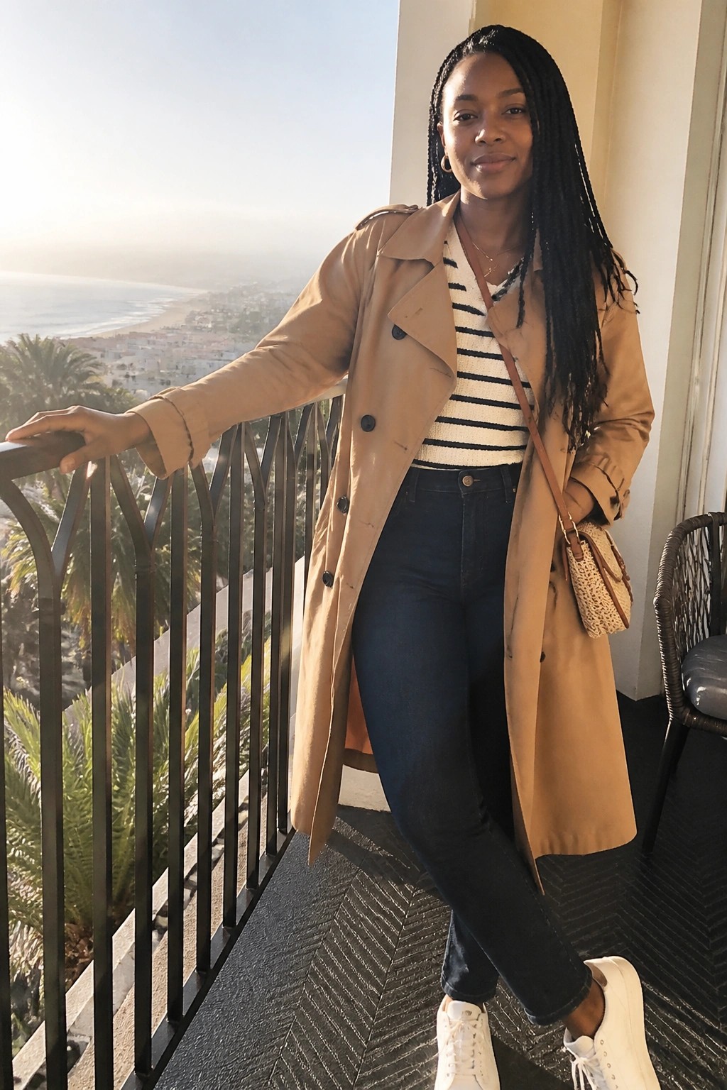 Woman in tan trench coat and jeans on balcony overlooking ocean at sunset