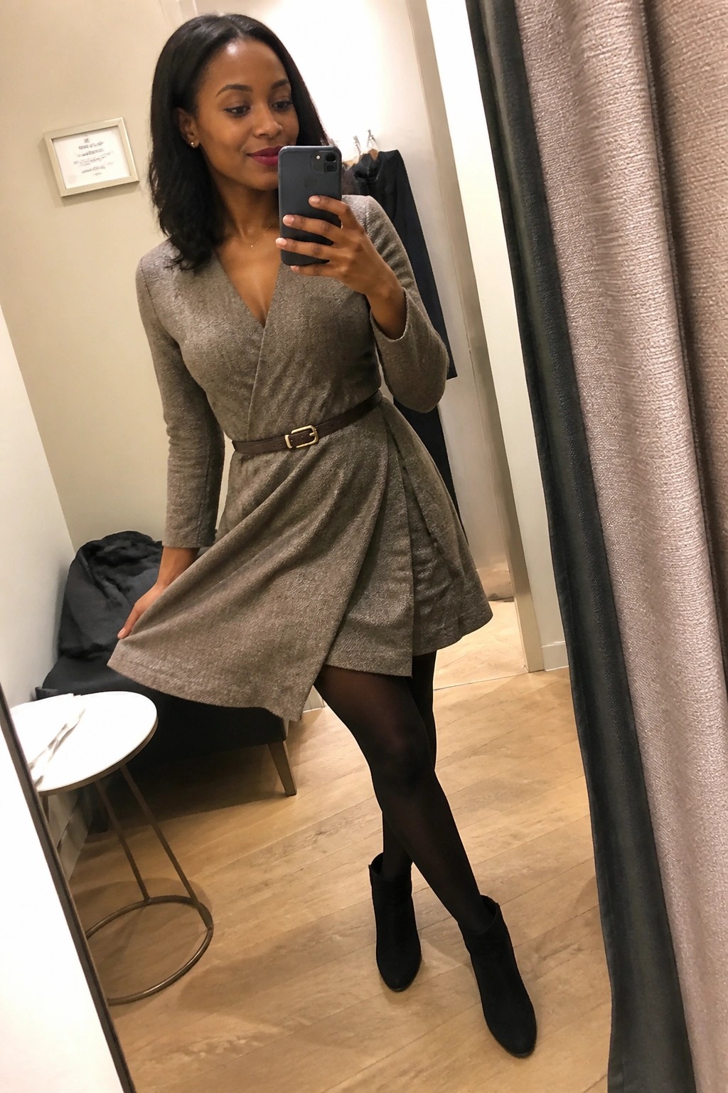 Woman in glittery taupe wrap dress and black boots taking mirror selfie in fitting room