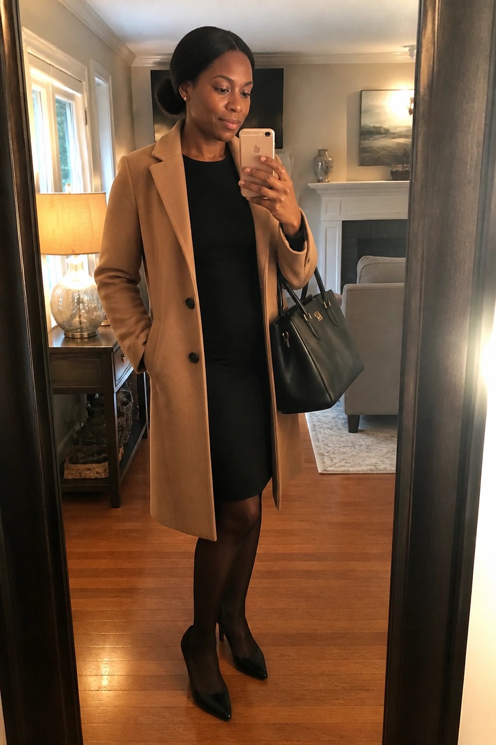 Woman wearing a camel wool coat over a black knee length dress with black tights black pointed heels and carrying a black leather handbag while holding a phone.