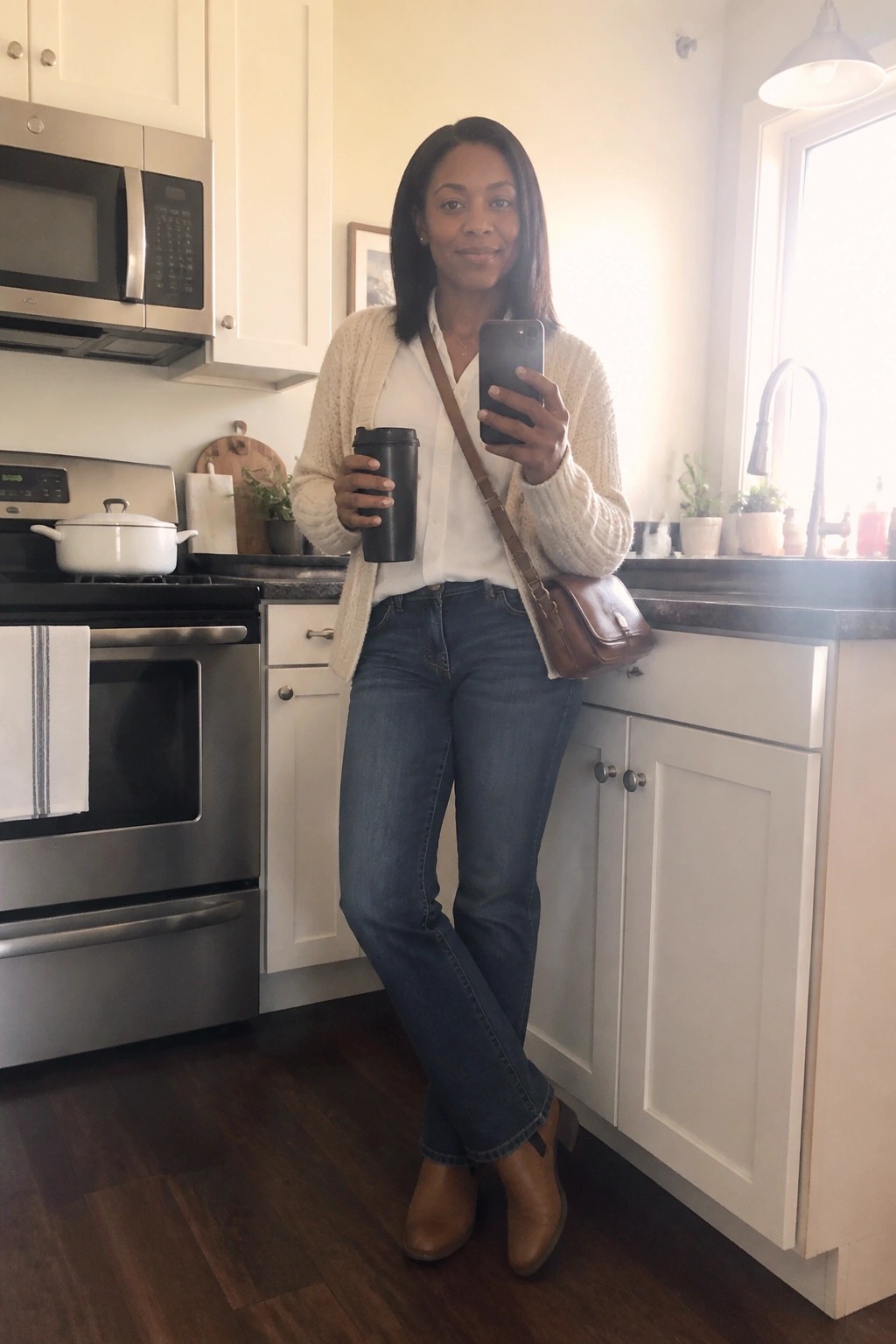 Woman in cream cardigan and jeans holding phone and coffee mug in kitchen