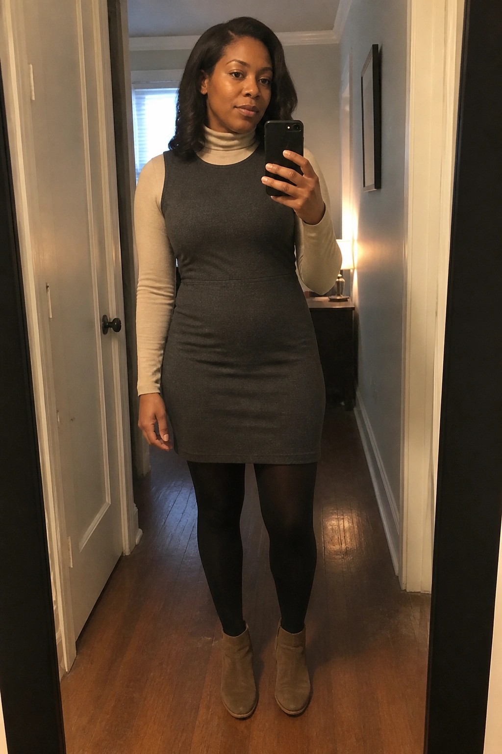 A woman wearing a gray sleeveless dress over a beige turtleneck, paired with black tights and taupe ankle boots, taking a mirror selfie in a hallway.