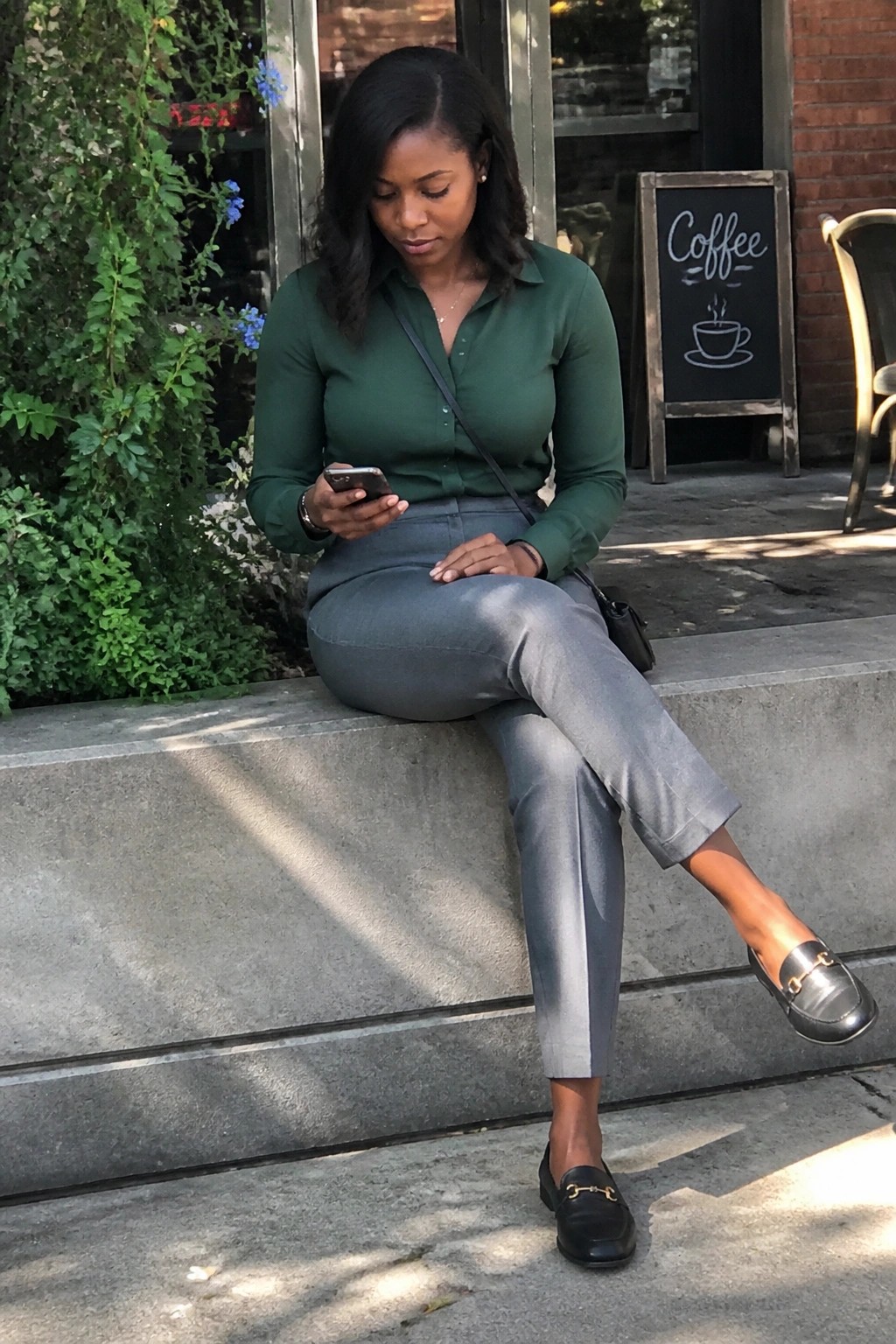 Woman in a green button-up shirt tucked into gray trousers paired with black loafers and subtle gold accessories, seated while viewing a phone.