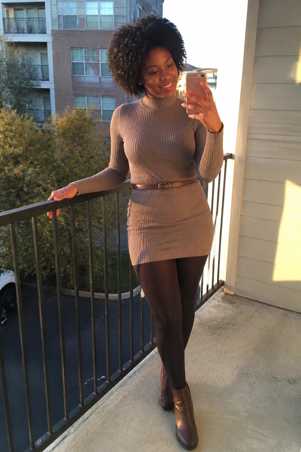 Woman with curly hair in beige sweater dress taking selfie on balcony