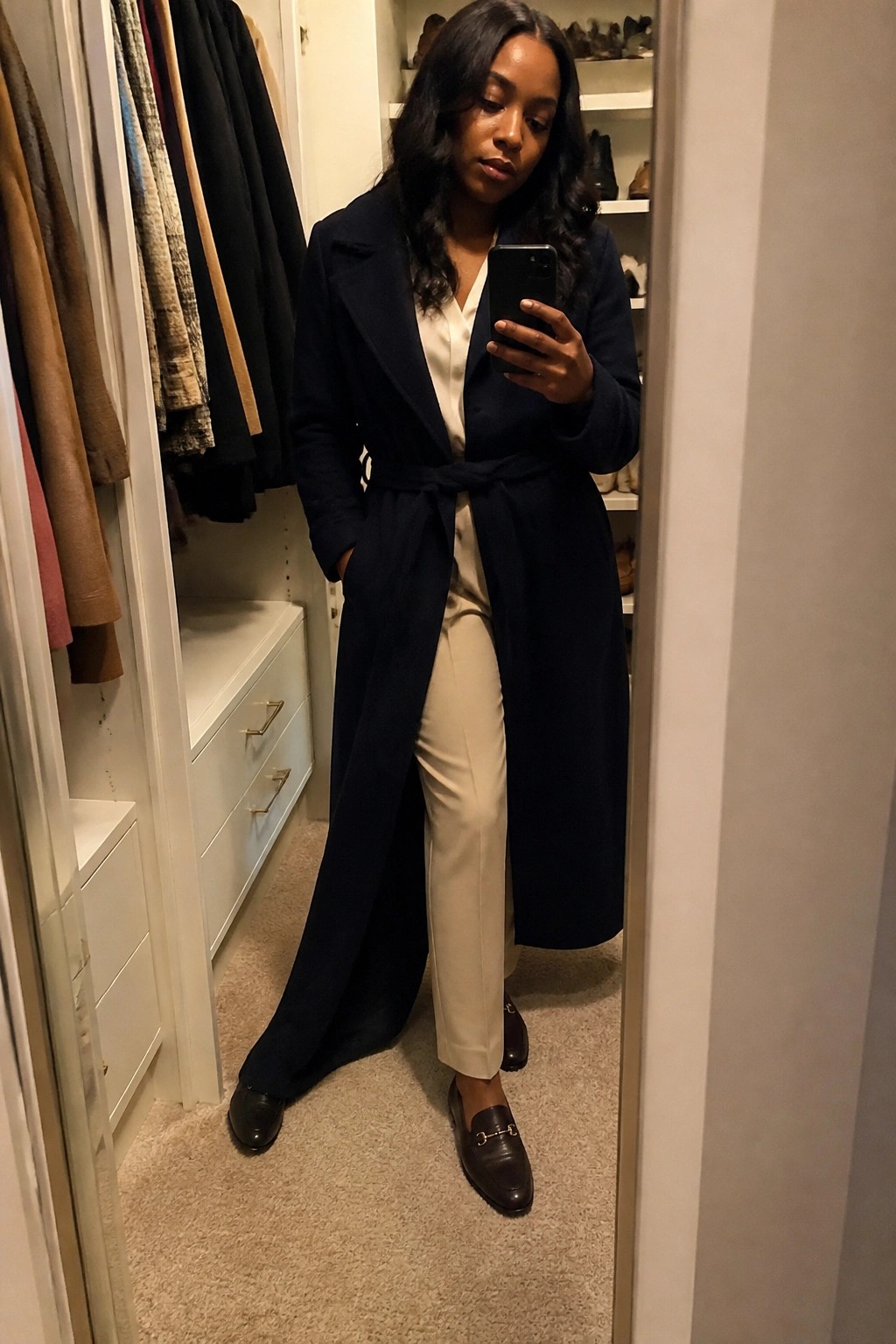 Woman in navy belted coat taking mirror selfie in walk-in closet