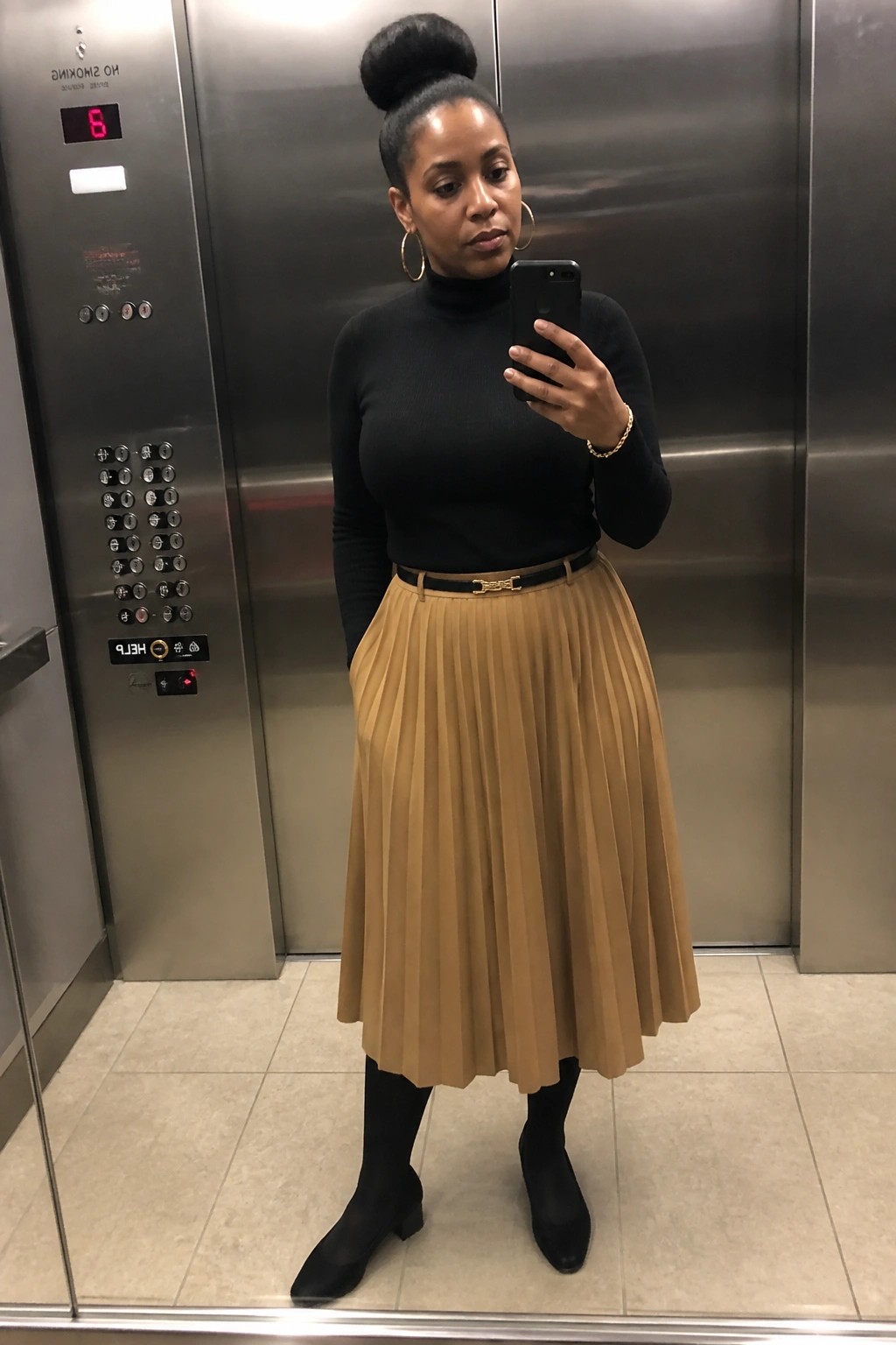 Woman in black turtleneck and tan pleated skirt taking selfie in elevator