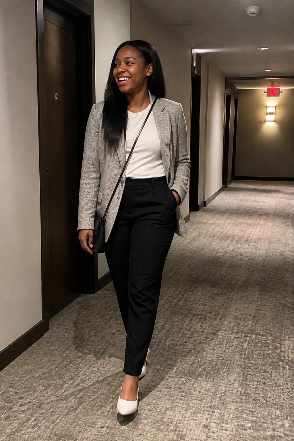 Black woman in gray tweed blazer over white tee black trousers and white pointed heels standing in hallway