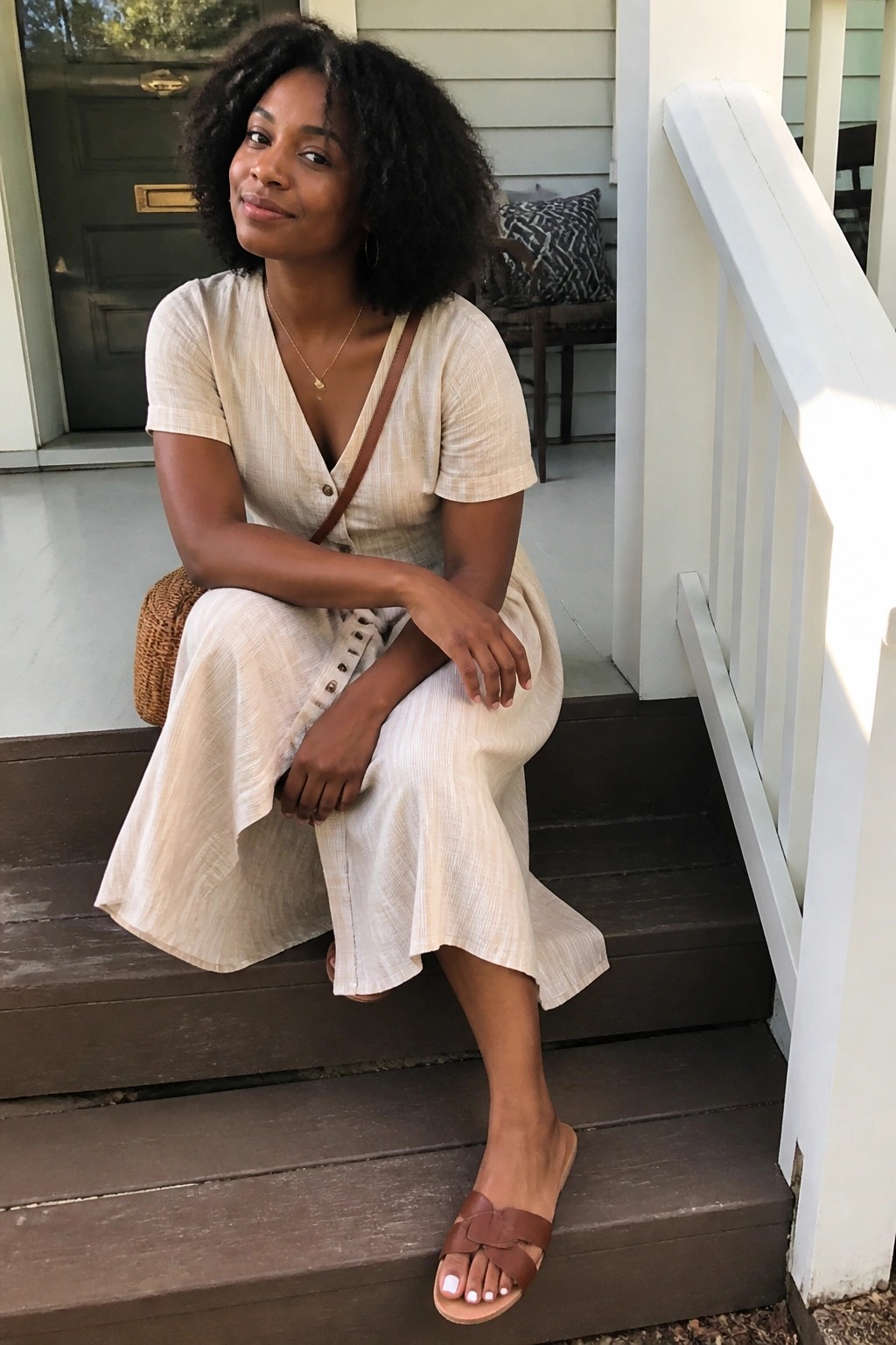 Black woman in cream linen midi dress with short sleeves, front buttons, brown crossbody strap, and brown leather slide sandals featuring a bow detail.