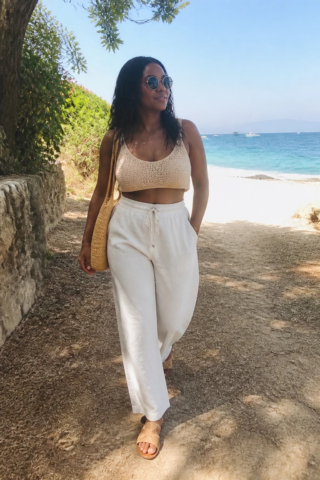 Woman in beige crochet crop top and white pants with straw bag on beach path
