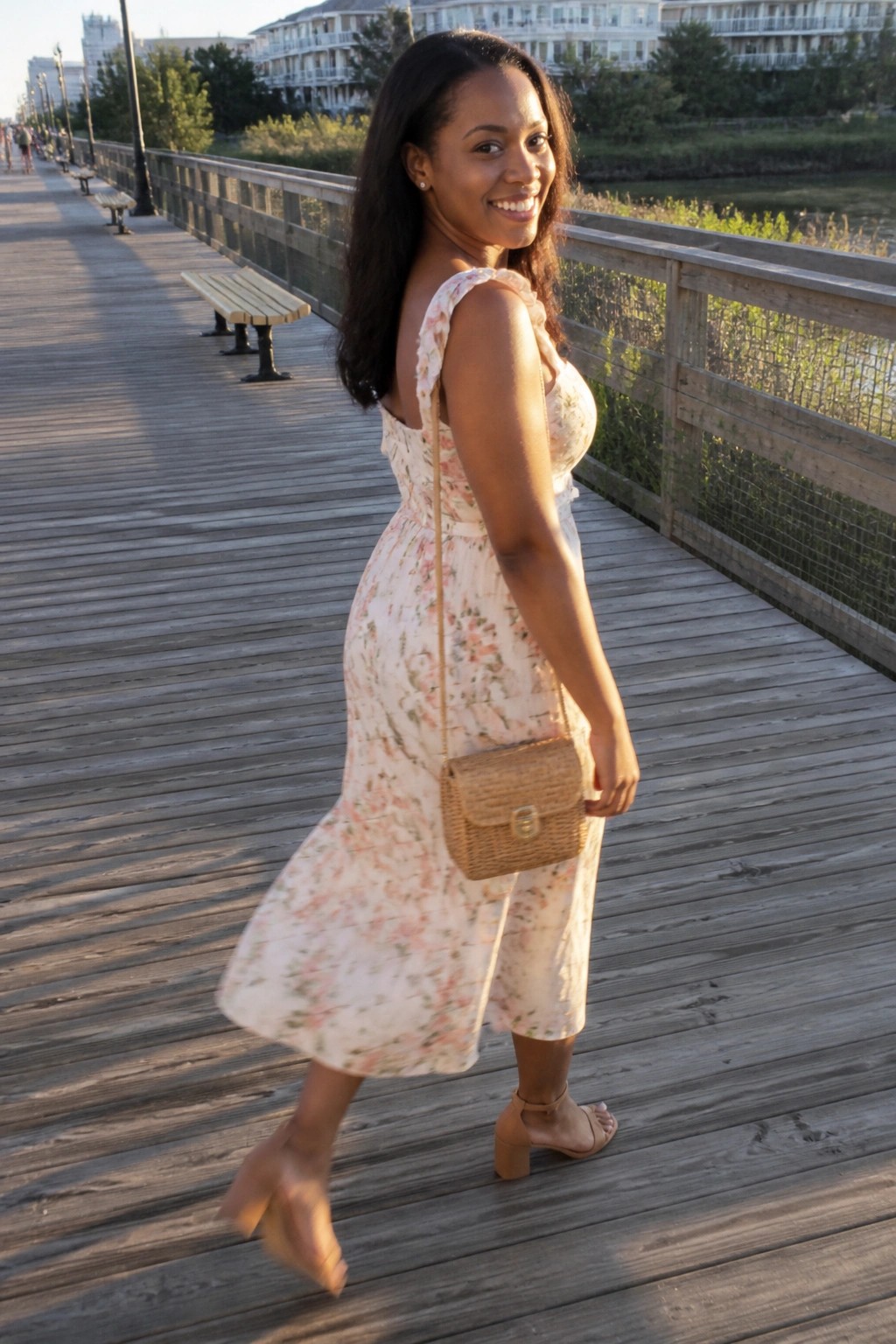 Woman in floral dress with woven purse smiling on wooden boardwalk at sunset
