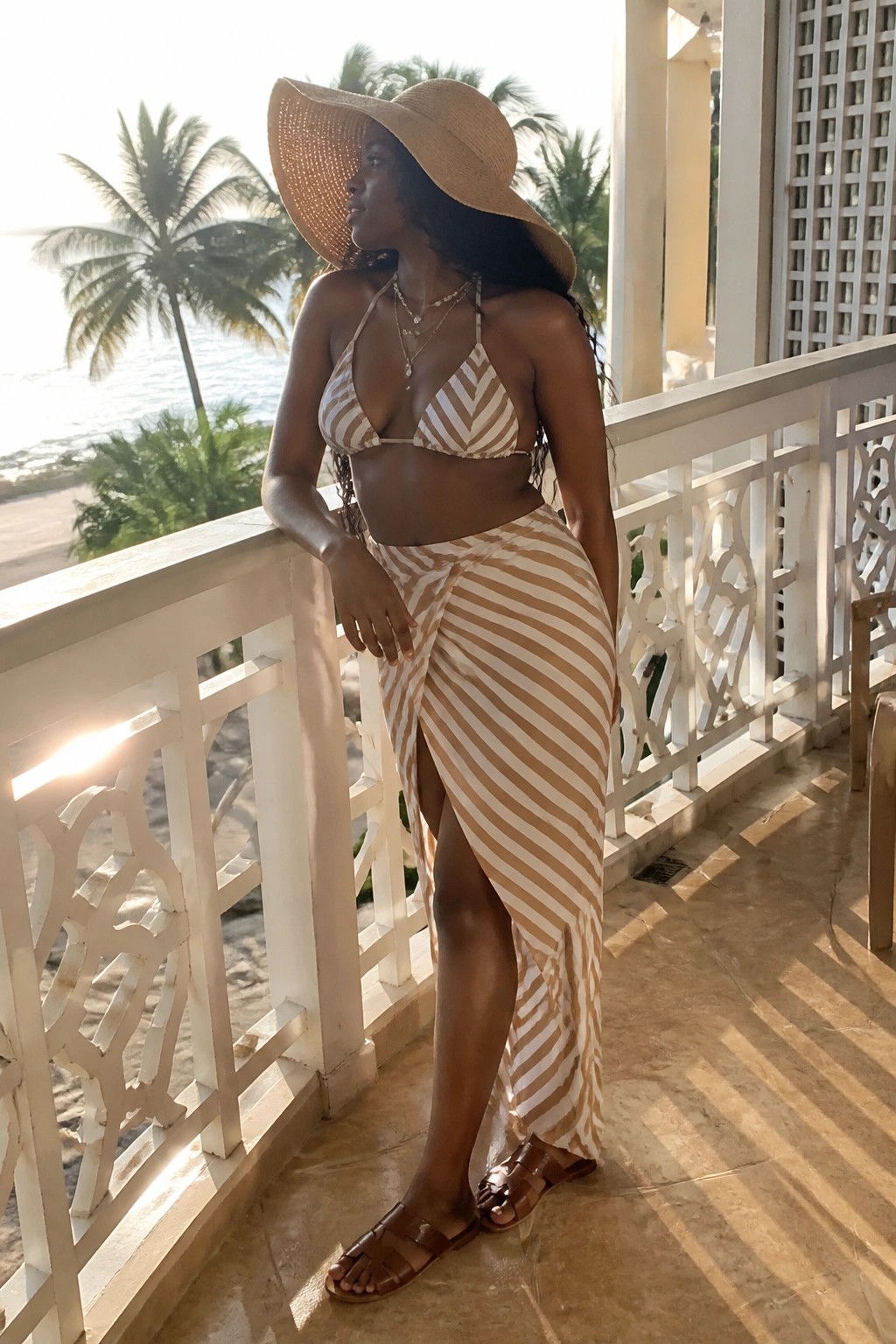 Woman in brown and white striped bikini top and matching striped sarong skirt with layered necklaces, straw hat, and brown slide sandals standing on a tiled balcony.