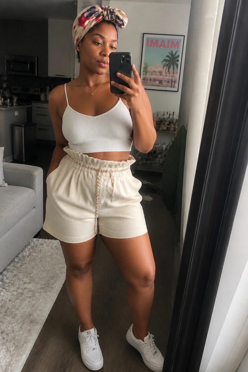 Woman in white crop top, cream shorts with chain belt, and headwrap taking mirror selfie