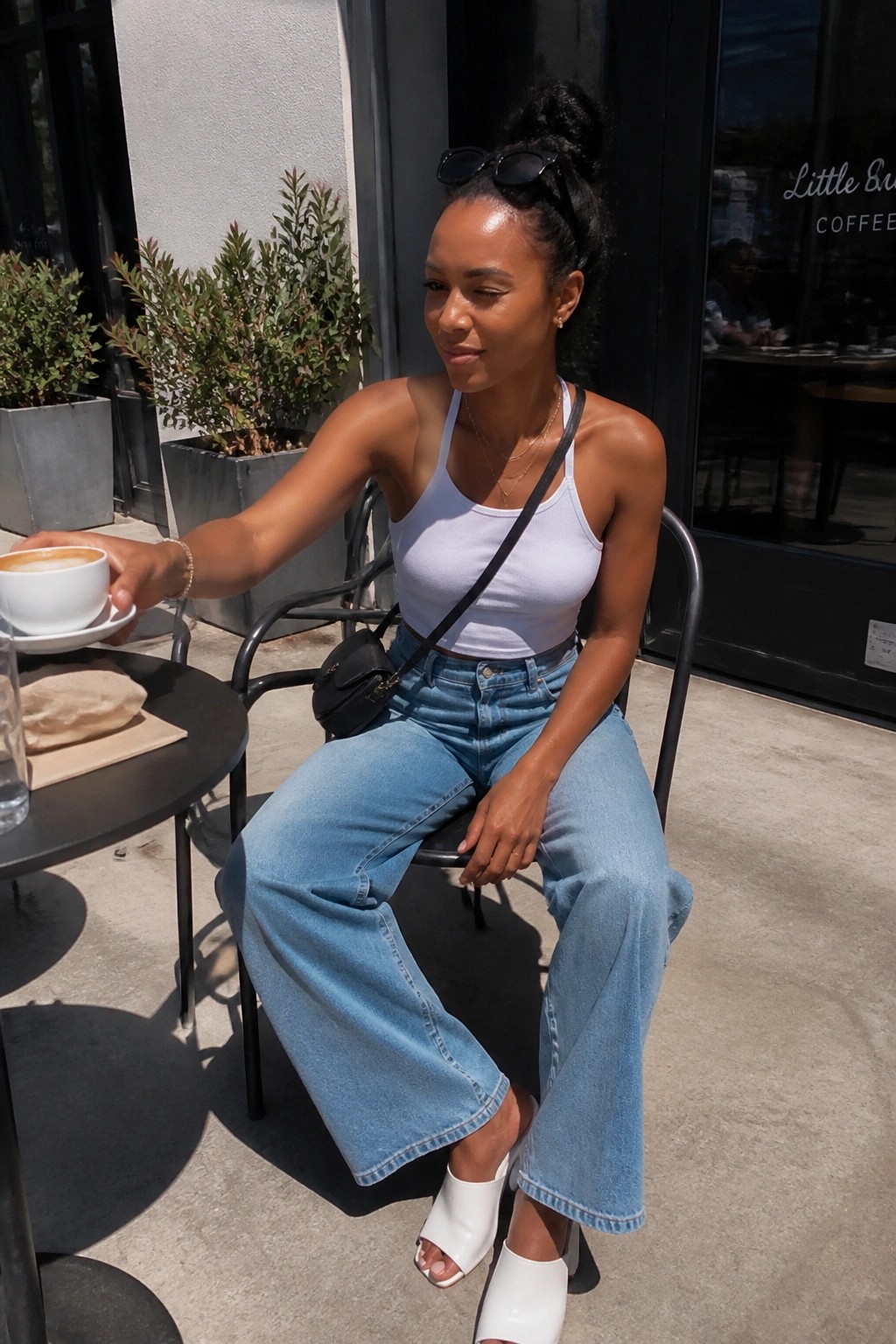 Woman in white tank top and blue wide-leg jeans holds coffee at sunny cafe.