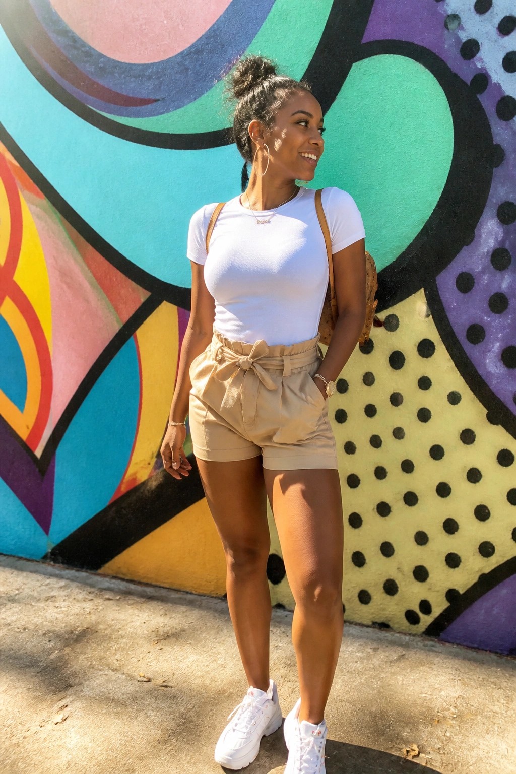 Woman in white top and beige tie-waist shorts smiling before vibrant mural