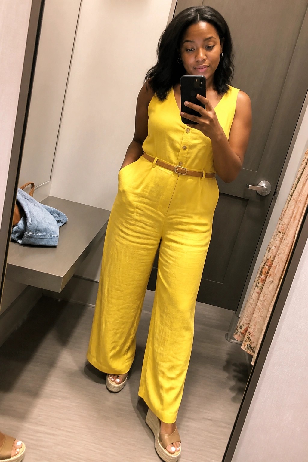 Woman wearing a bright yellow sleeveless linen jumpsuit with front buttons, brown belt, wide legs, and tan wedge espadrille sandals while taking a mirror selfie.