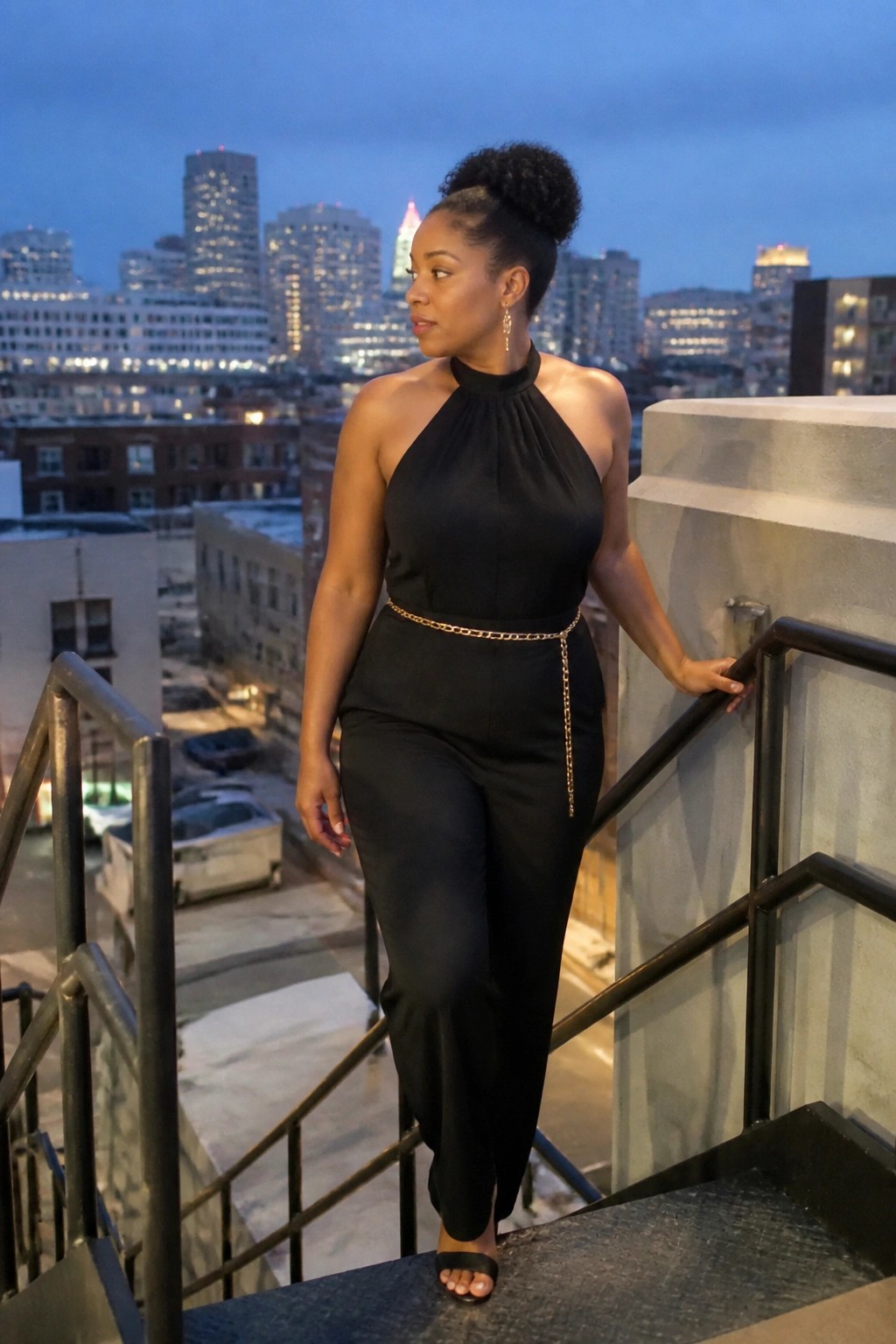 Woman in black halter jumpsuit with gold chain belt on city stairs at night