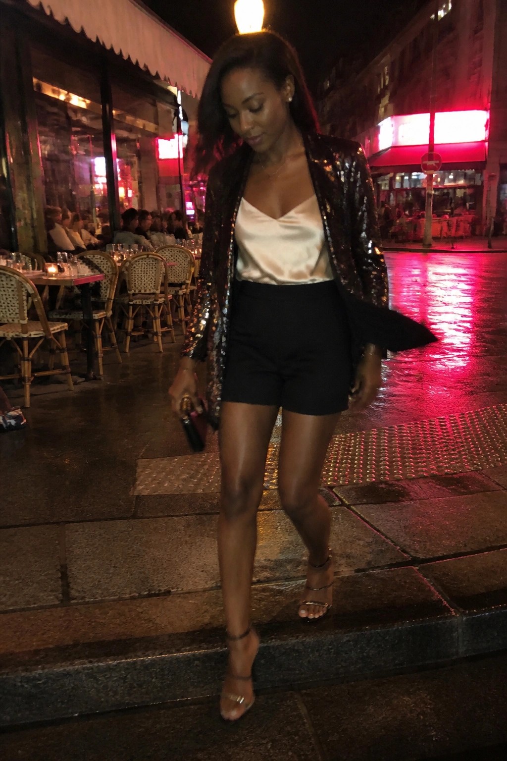Woman in sequined jacket and black shorts on wet city street at night.