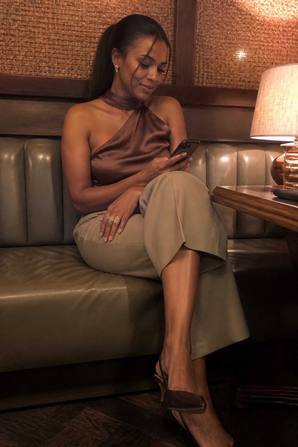 Woman in brown satin top and green pants checks phone on leather booth.