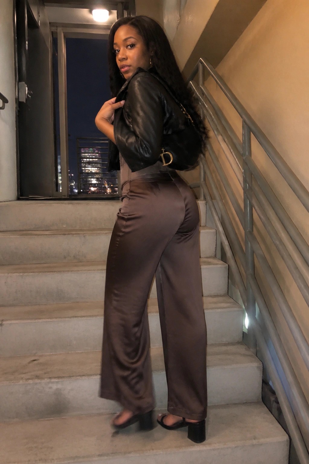 Woman in black leather jacket and brown satin pants on nighttime stairs