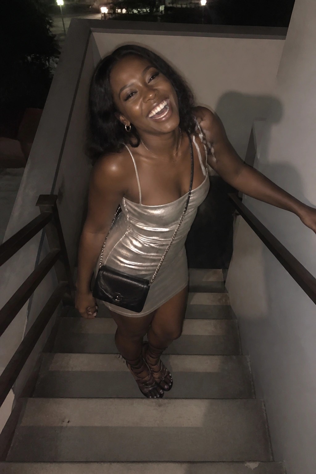 Woman in short metallic silver mini dress with thin straps, black chain-strap crossbody bag, and strappy heels standing on concrete stairs.