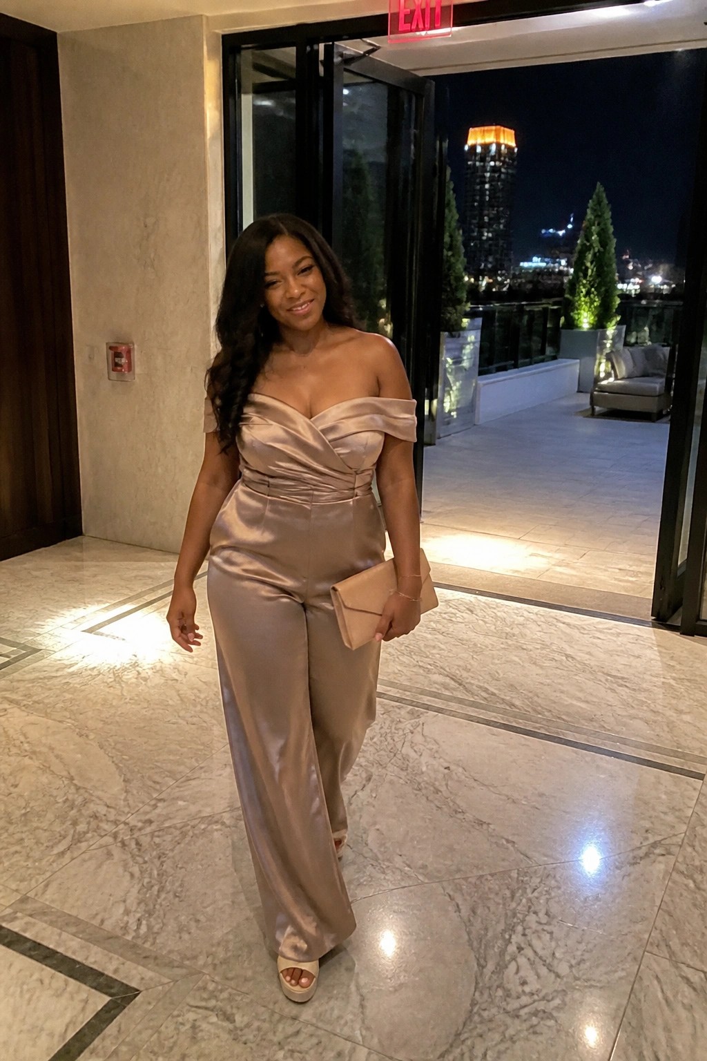 Woman in beige satin off-shoulder jumpsuit holding clutch near open exit door at night.