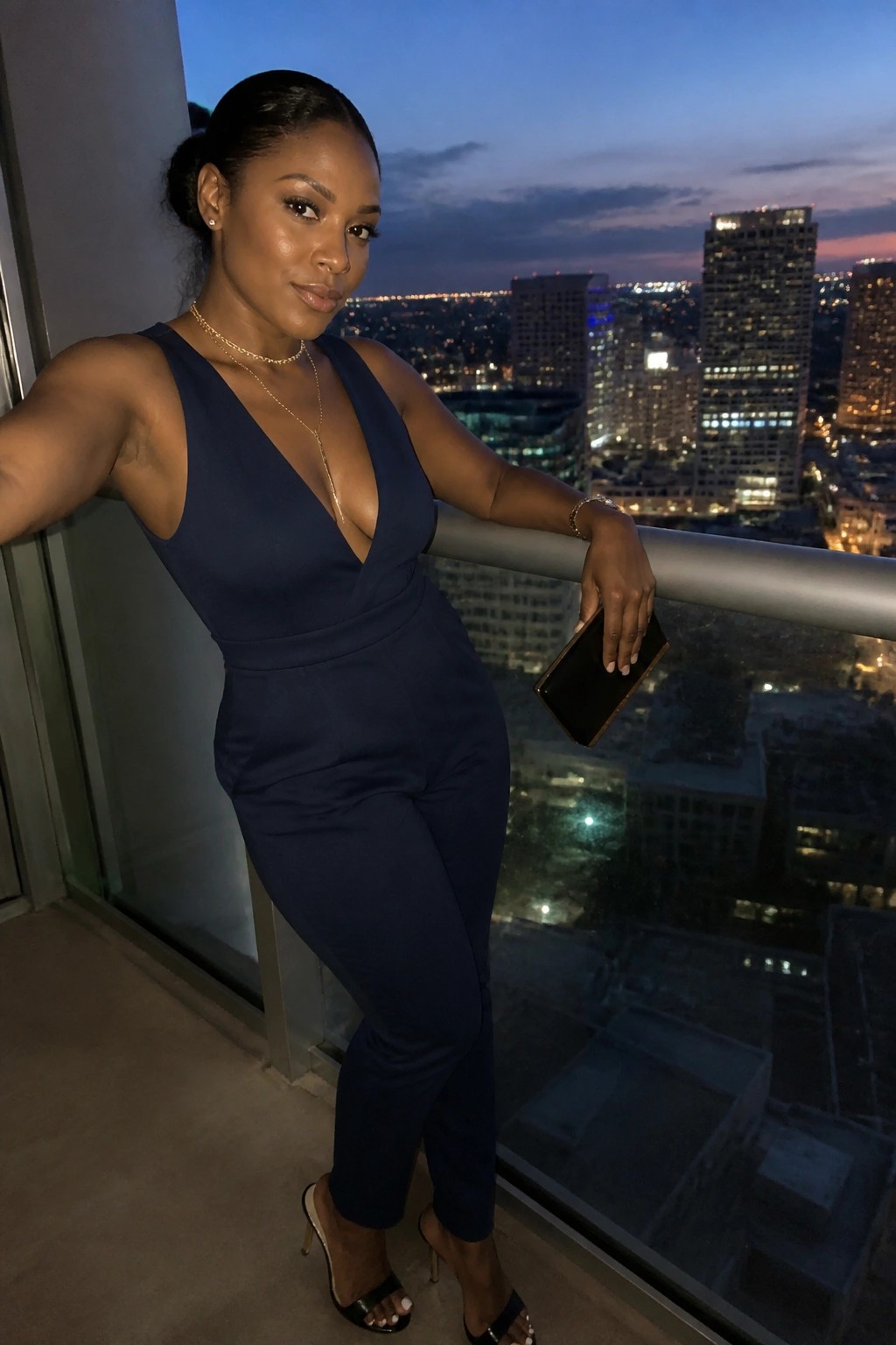 Woman in navy jumpsuit leaning on balcony railing above glowing city at dusk