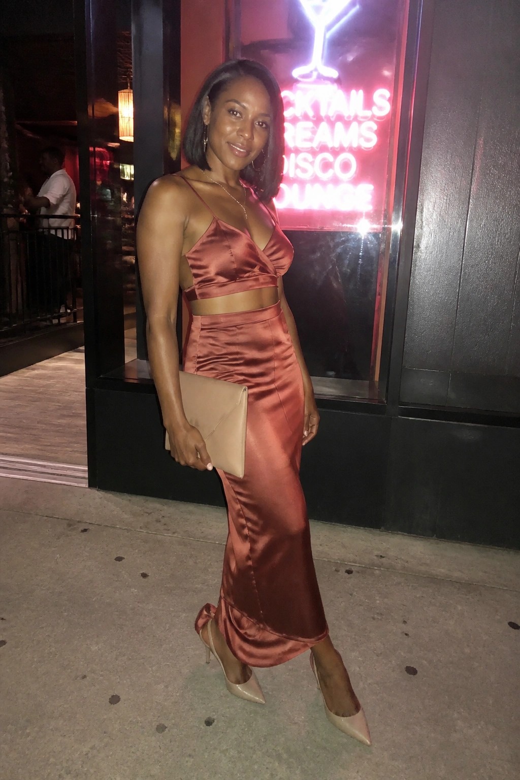 Woman in rose satin crop top and skirt holding beige clutch by neon lounge sign