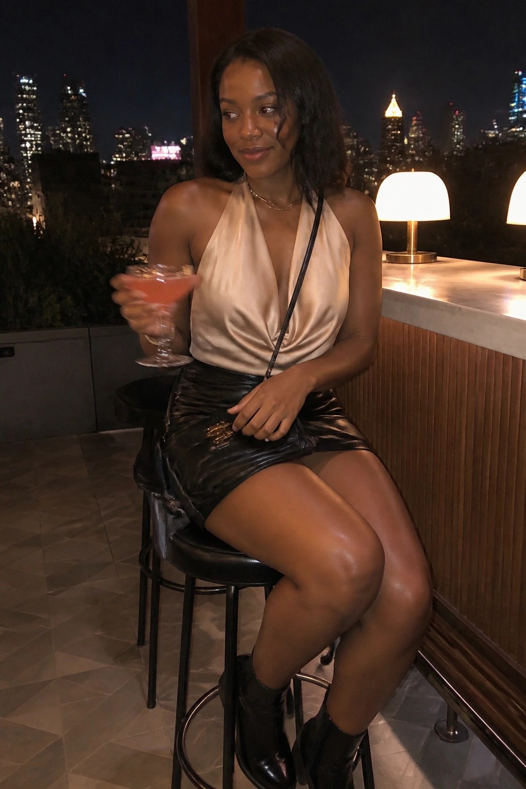 Woman in beige halter top and black leather skirt holding pink cocktail at night bar