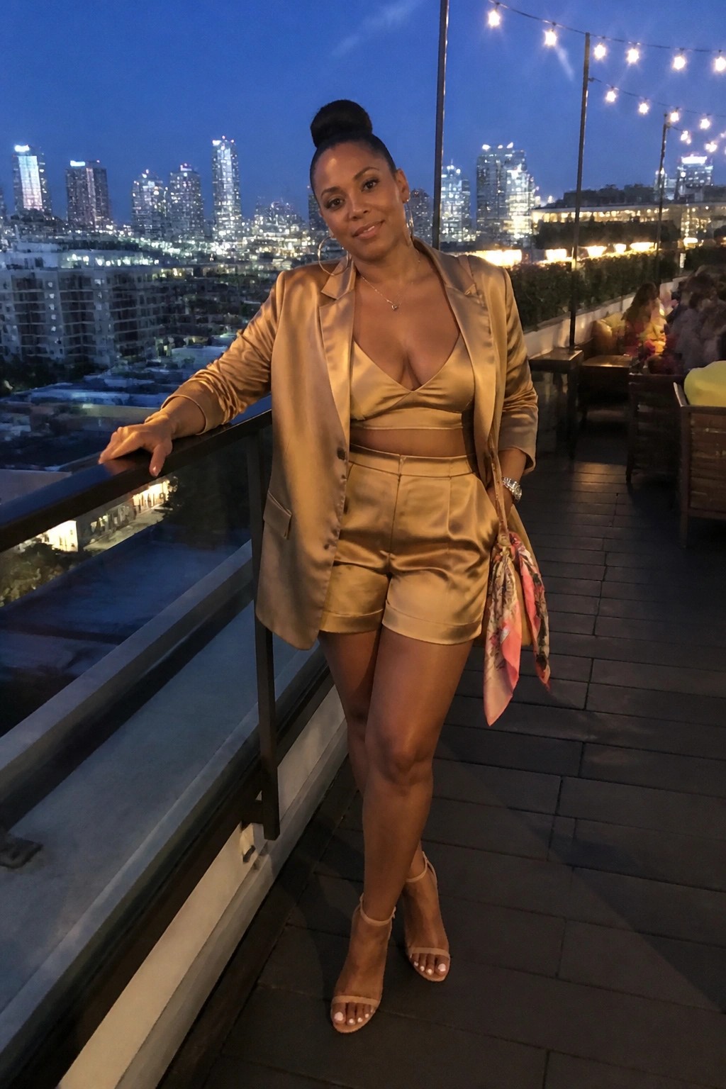 Woman in gold satin blazer and shorts on rooftop overlooking city lights at night.