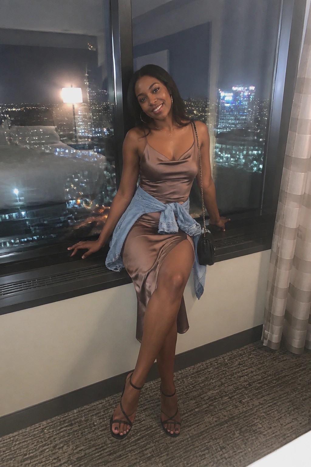 Smiling woman in satin dress with tied denim jacket by nighttime city window