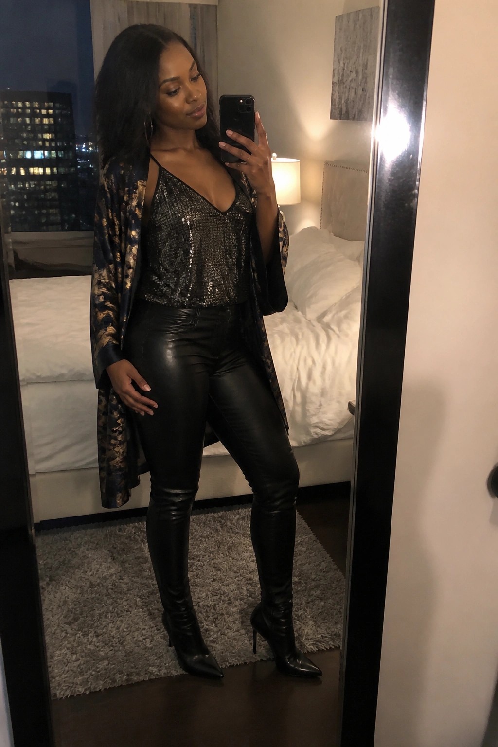 Woman in sequined top and leather pants taking mirror selfie in bedroom