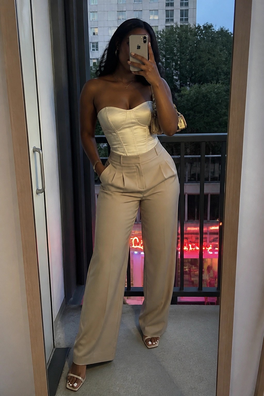 Woman in beige corset top and wide-leg pants taking mirror selfie on balcony