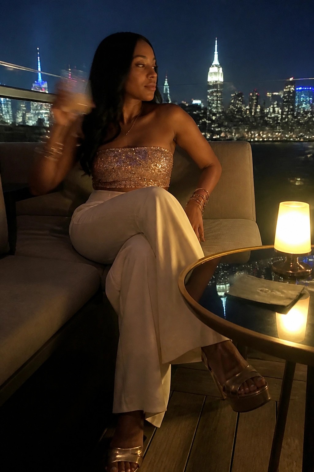 Woman in sequined top and white pants posing before illuminated NYC skyline at night.