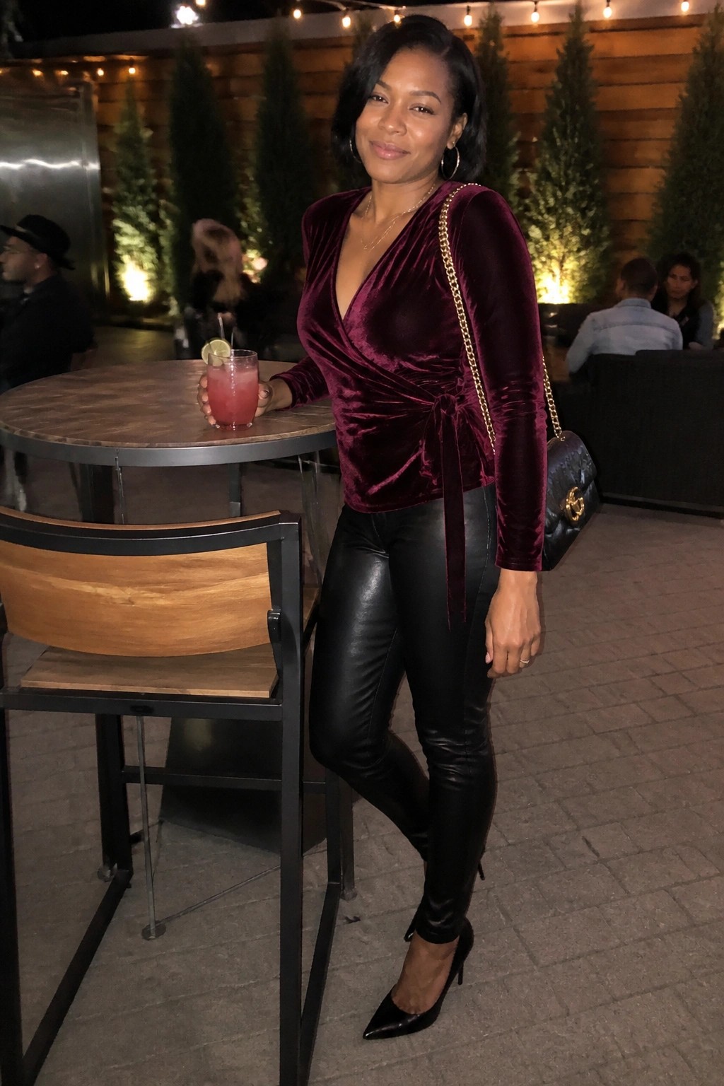 Woman in burgundy velvet top and black leather pants at outdoor bar table with drink