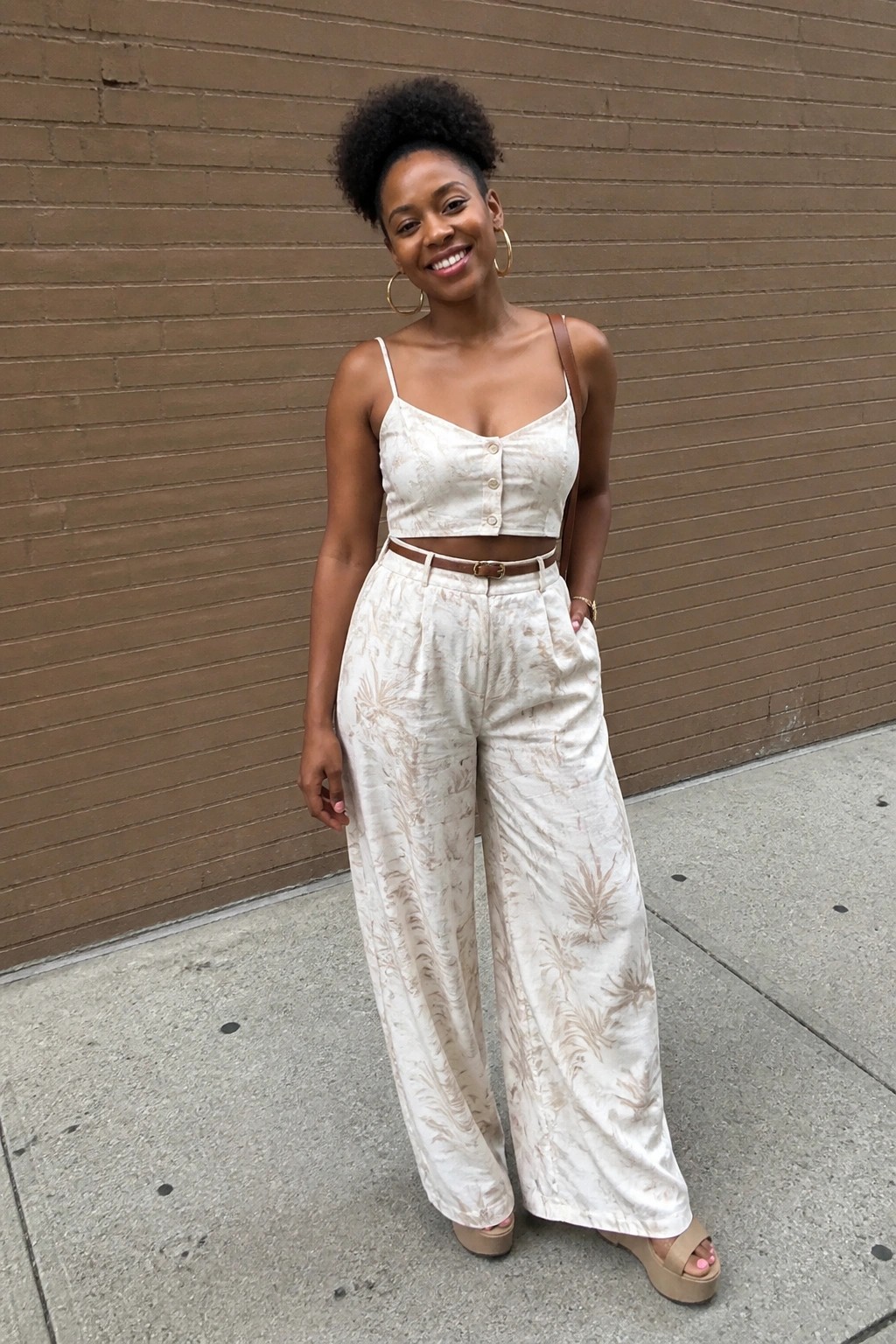 Smiling woman in patterned cream crop top and wide-leg pants by brick wall