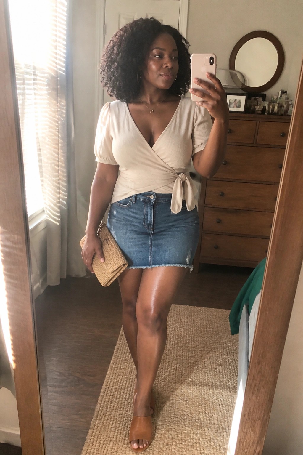 Woman in beige wrap top and denim mini skirt taking mirror selfie with woven clutch