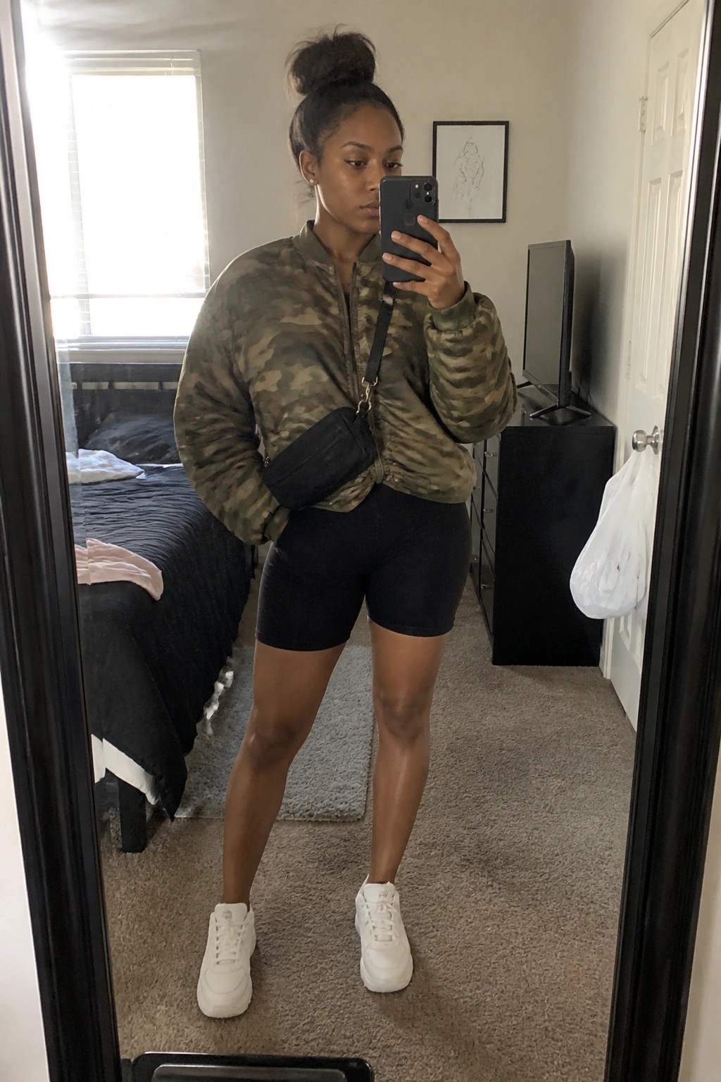 Woman in camouflage jacket and black shorts taking mirror selfie in bedroom