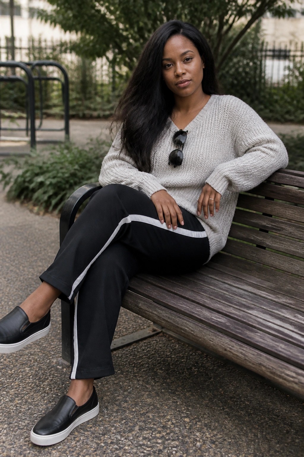 Woman seated on a bench wearing an oversized light gray chunky knit V-neck sweater with black pants featuring white side stripes and black slip-on sneakers with white soles, sunglasses tucked into the sweater neckline.