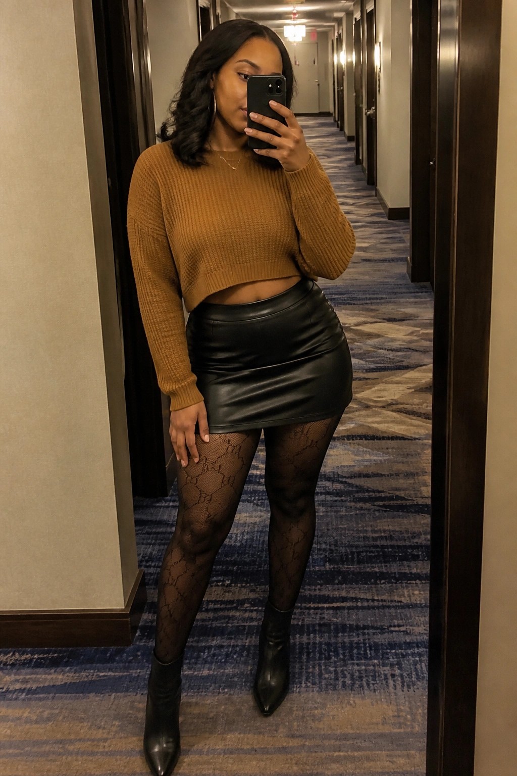 Woman in brown cropped ribbed knit sweater, black leather mini skirt, black GG patterned tights, and pointed black ankle boots taking a mirror selfie in a hallway.