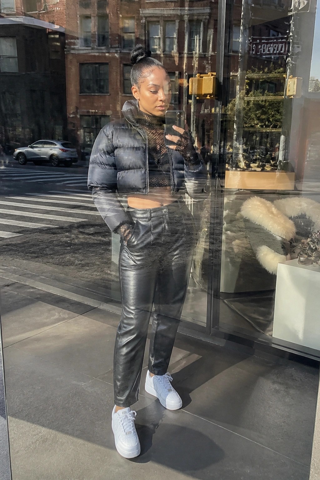 Woman in black puffer jacket and leather pants taking mirror selfie on city street.