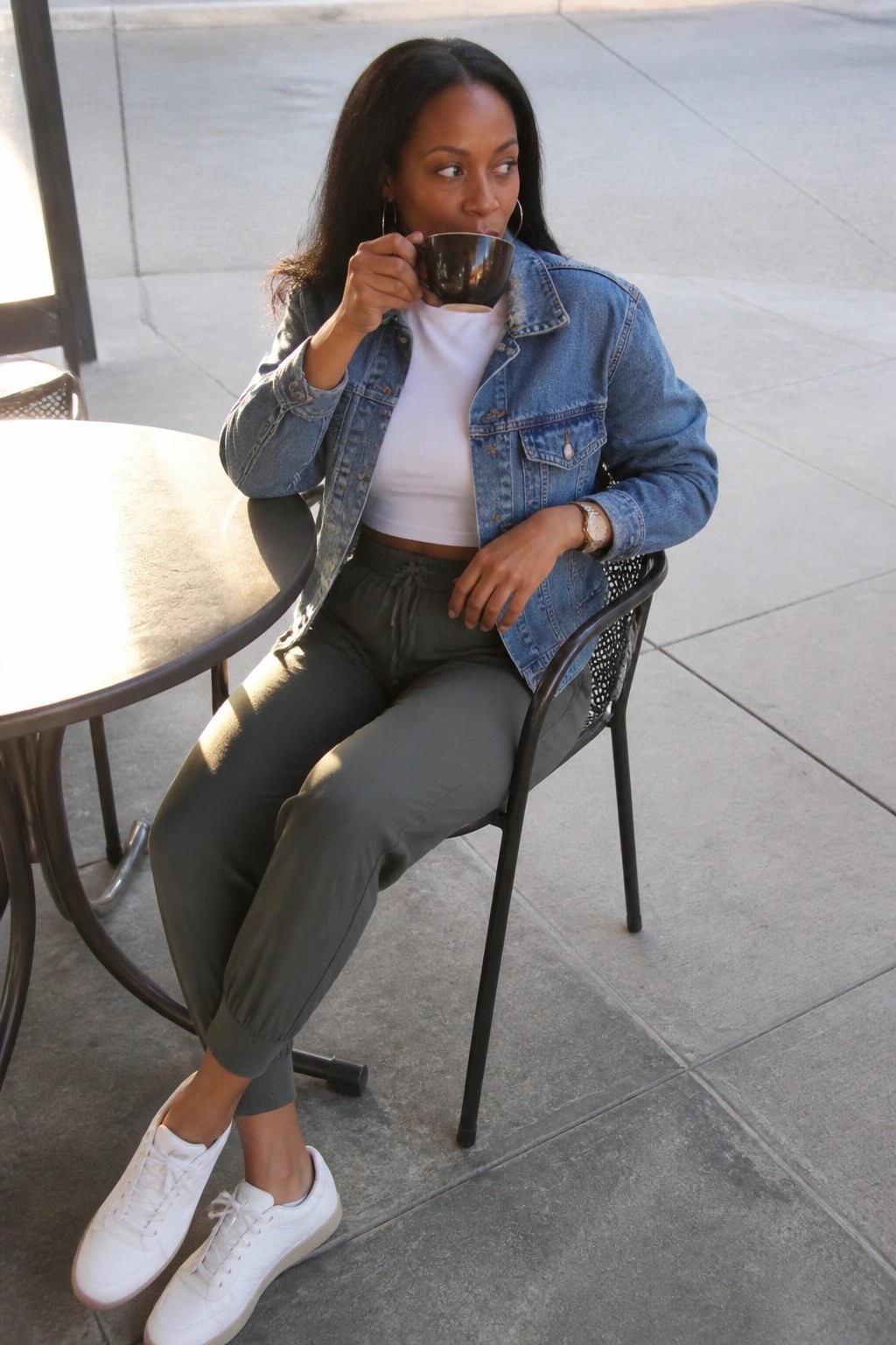 Woman in denim jacket sips coffee at outdoor sidewalk cafe table