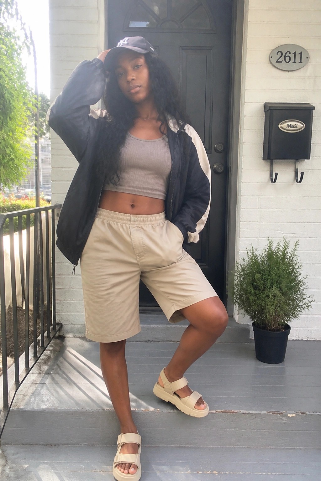 Woman in beige shorts, crop top, and black jacket posing on porch steps.