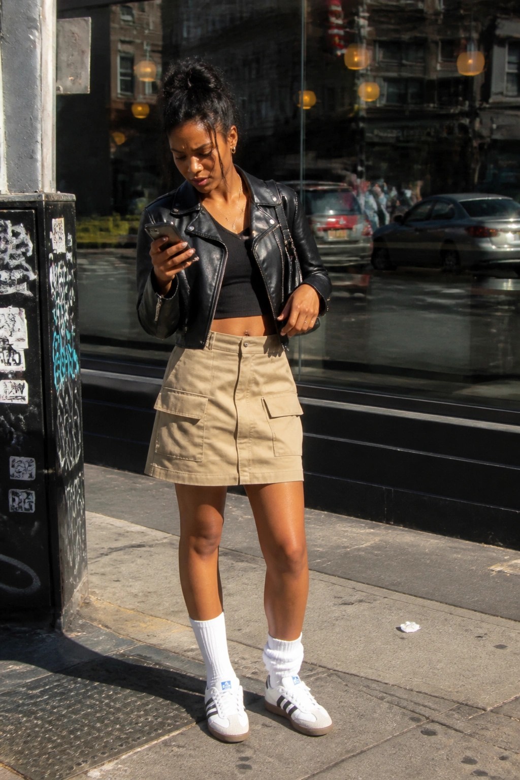 Woman in black leather jacket and beige cargo skirt checks phone on sidewalk
