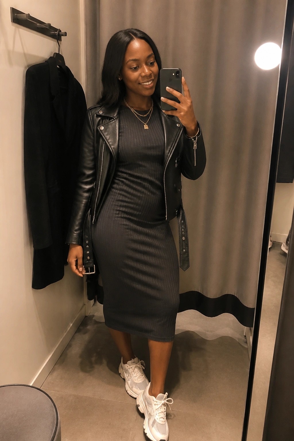 Woman in black leather jacket and gray dress taking mirror selfie in fitting room