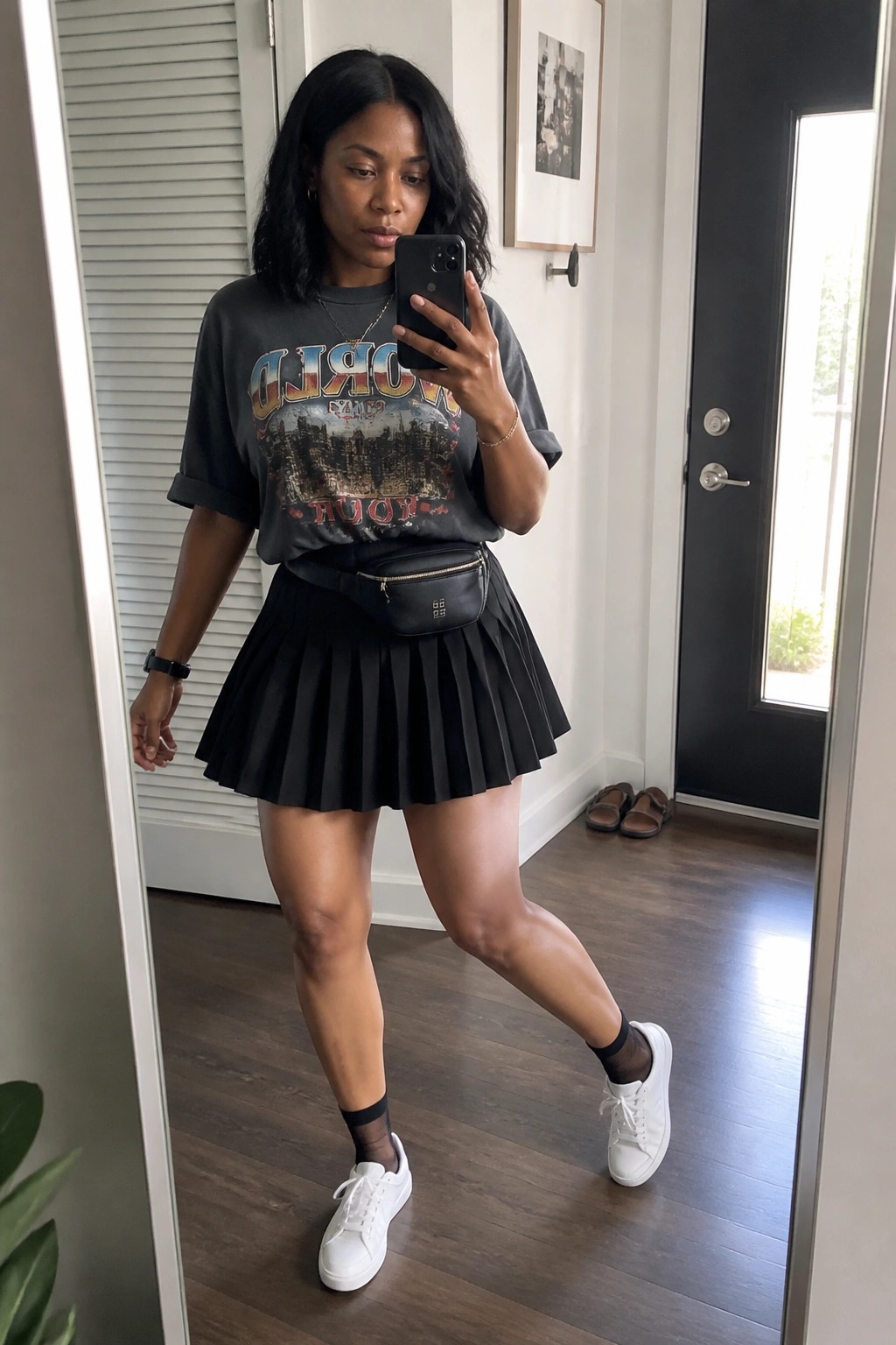 Woman in black graphic tee and pleated skirt taking mirror selfie wearing white sneakers