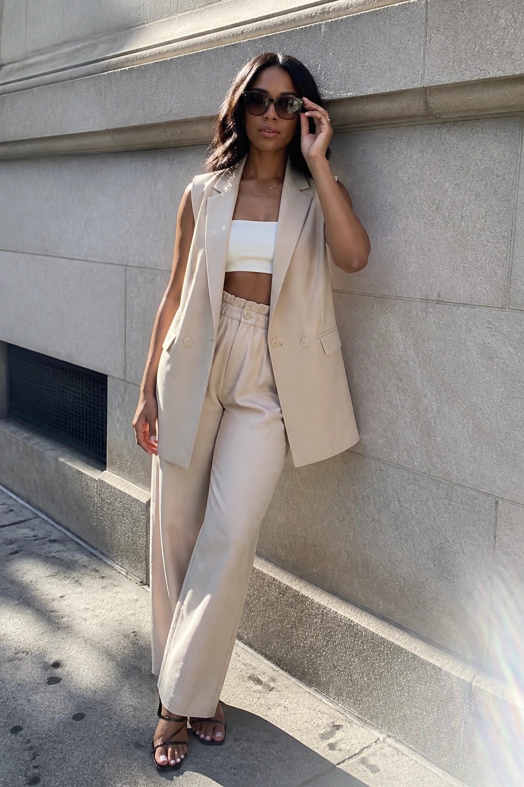 Woman in beige sleeveless blazer and wide-leg pants posing by stone wall