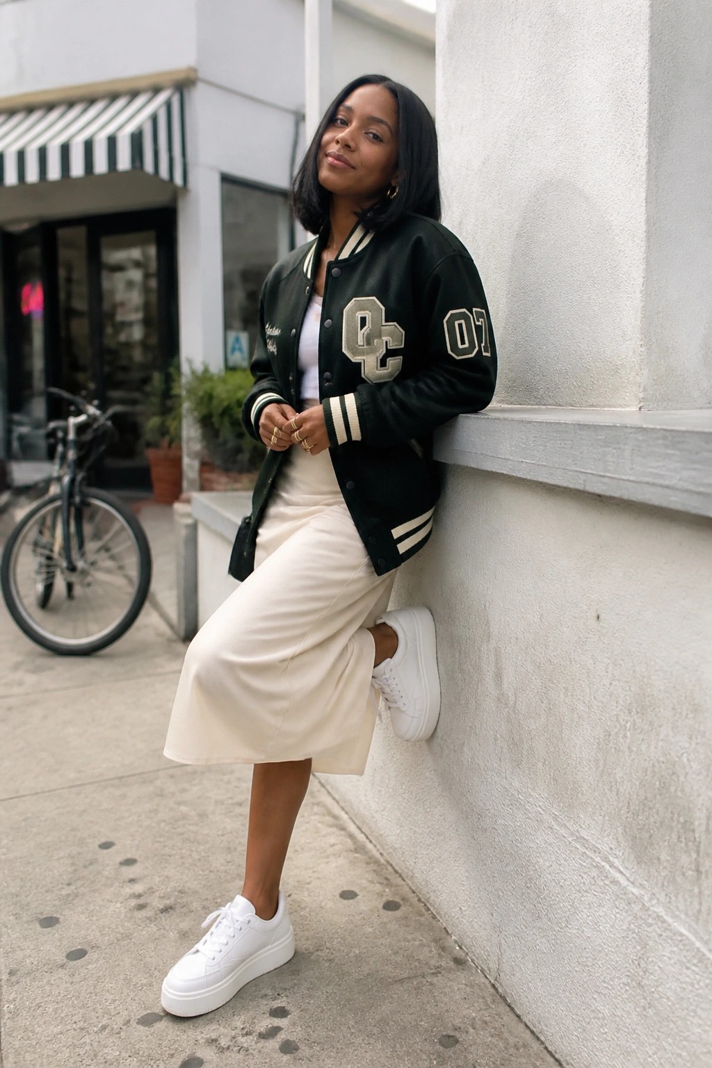 Woman in black varsity jacket and cream skirt leaning on urban stone wall.